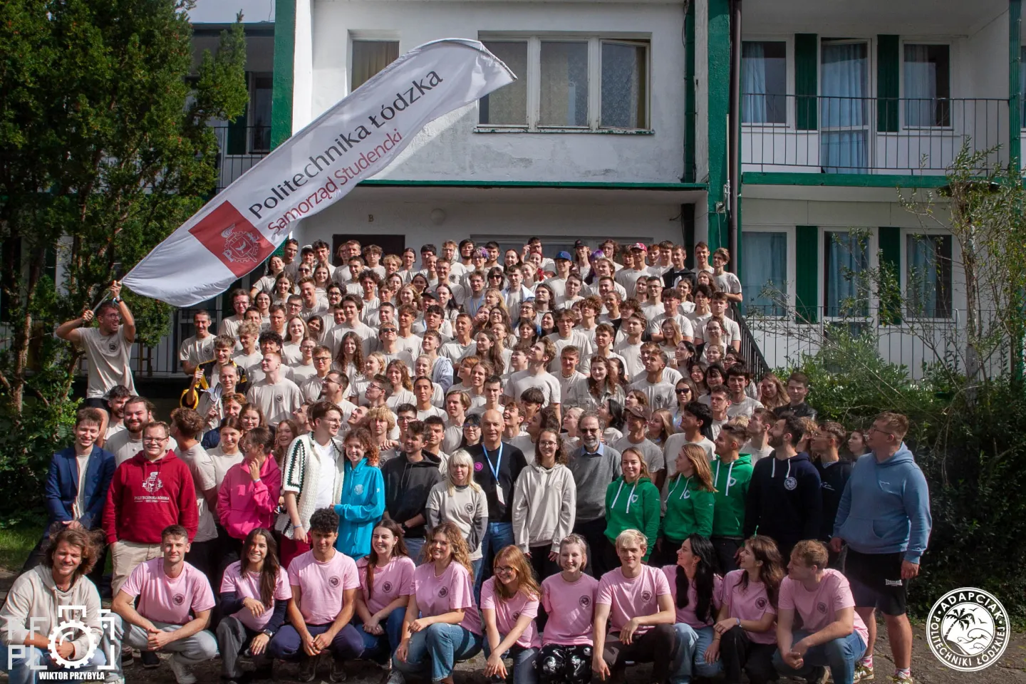 Duża grupa studentów stoi przed wielopiętrowym budynkiem. Większość osób ma na sobie beżowe koszulki, a kilka w pierwszym rzędzie ubrania w intensywnych kolorach. Po lewej stronie widoczny jest baner Samorządu Studenckiego PŁ. Zdjęcie przedstawia wspólnotę studentów zgromadzonych do pamiątkowej fotografii.