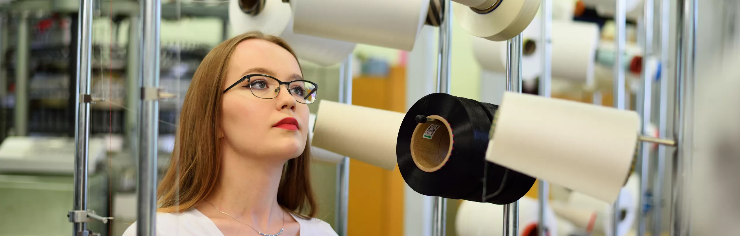 Kobieta stoi przy szpulach nici w hali produkcyjnej.